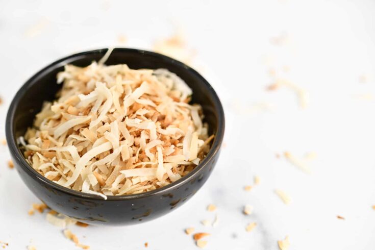Toasted coconut in a small black bowl on a marble counter with toasted coconut scattered across the surface.