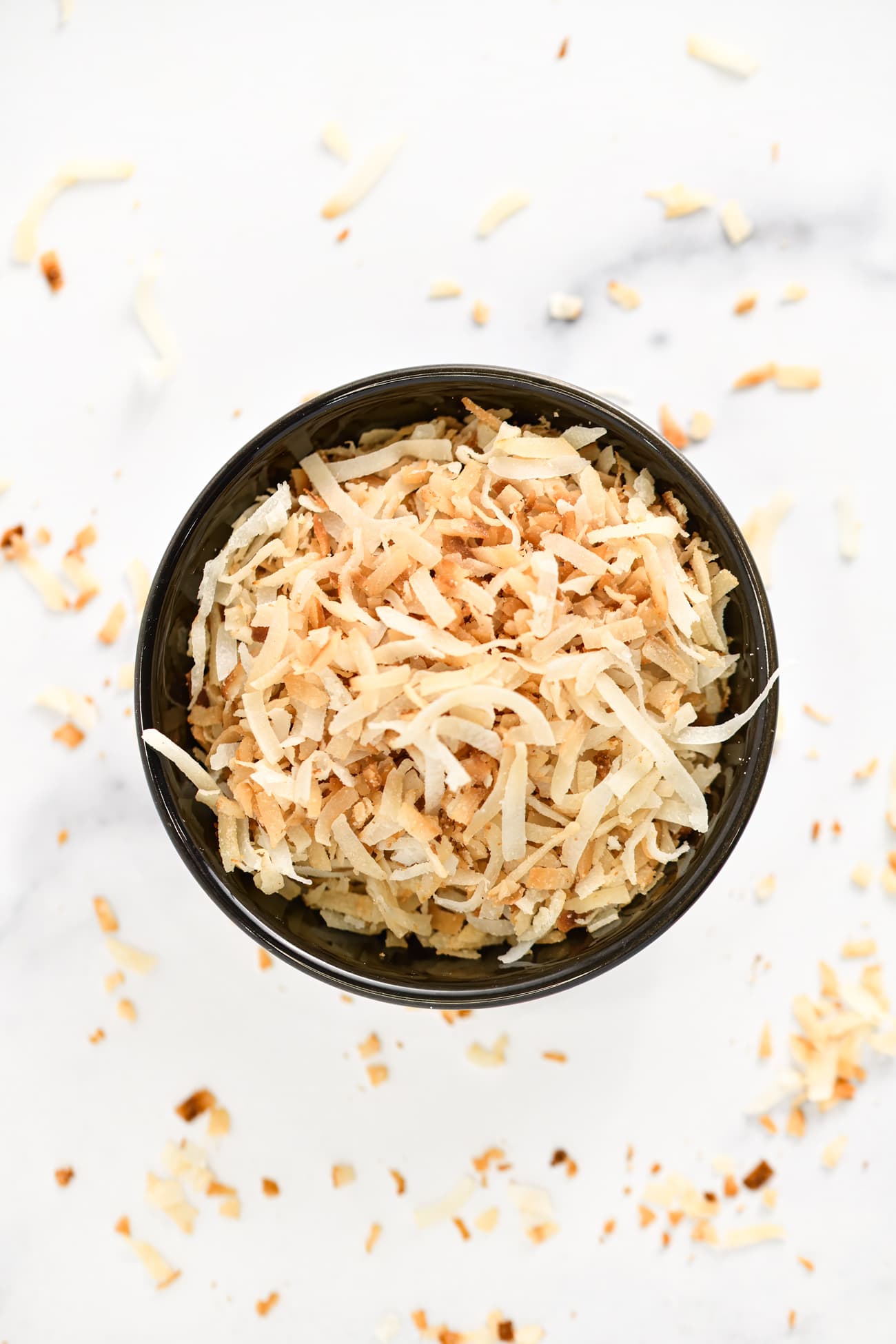 Overhead view of toasted coconut scattered on a marble counter with a small black bowl filled with more toasted coconut in the center