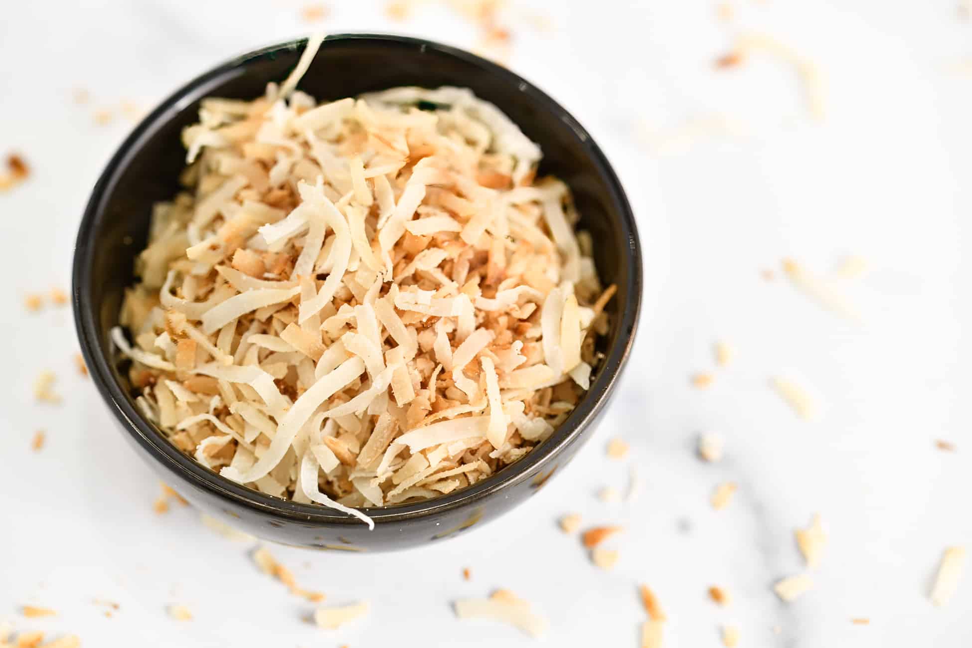 Toasted coconut in a small black bowl on a marble counter with toasted coconut scattered across the surface.