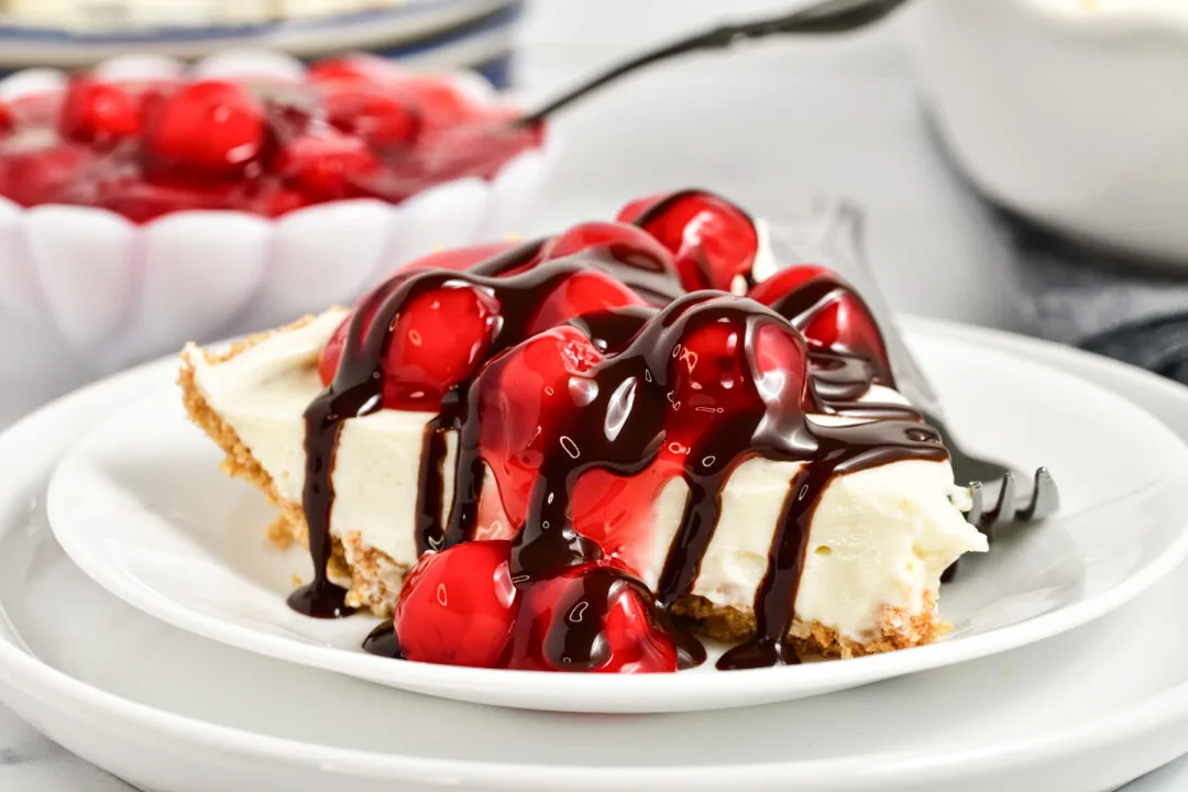 A slice of cheesecake topped with glossy red cherries and drizzled with chocolate sauce is placed on a white plate. In the background, a bowl of cherry topping is partially visible. A fork rests on the edge of the plate.