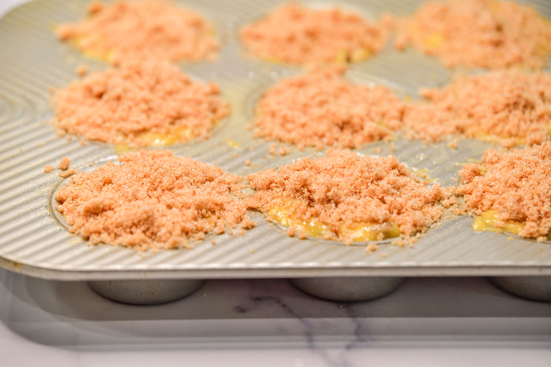 Silver muffin tin filled with muffin batter and streusel topping on top ready to go into the oven. 