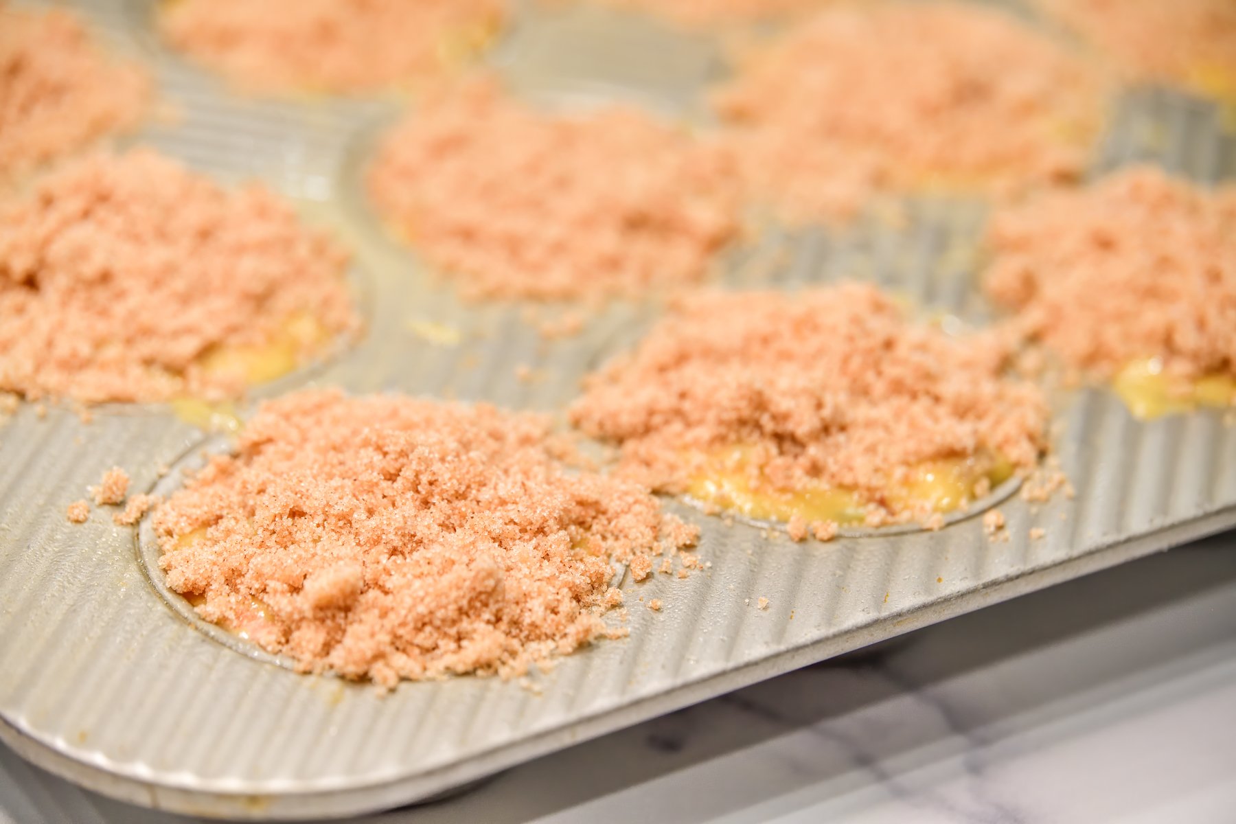 Silver muffin tin filled with muffin batter and streusel topping on top ready to go into the oven.