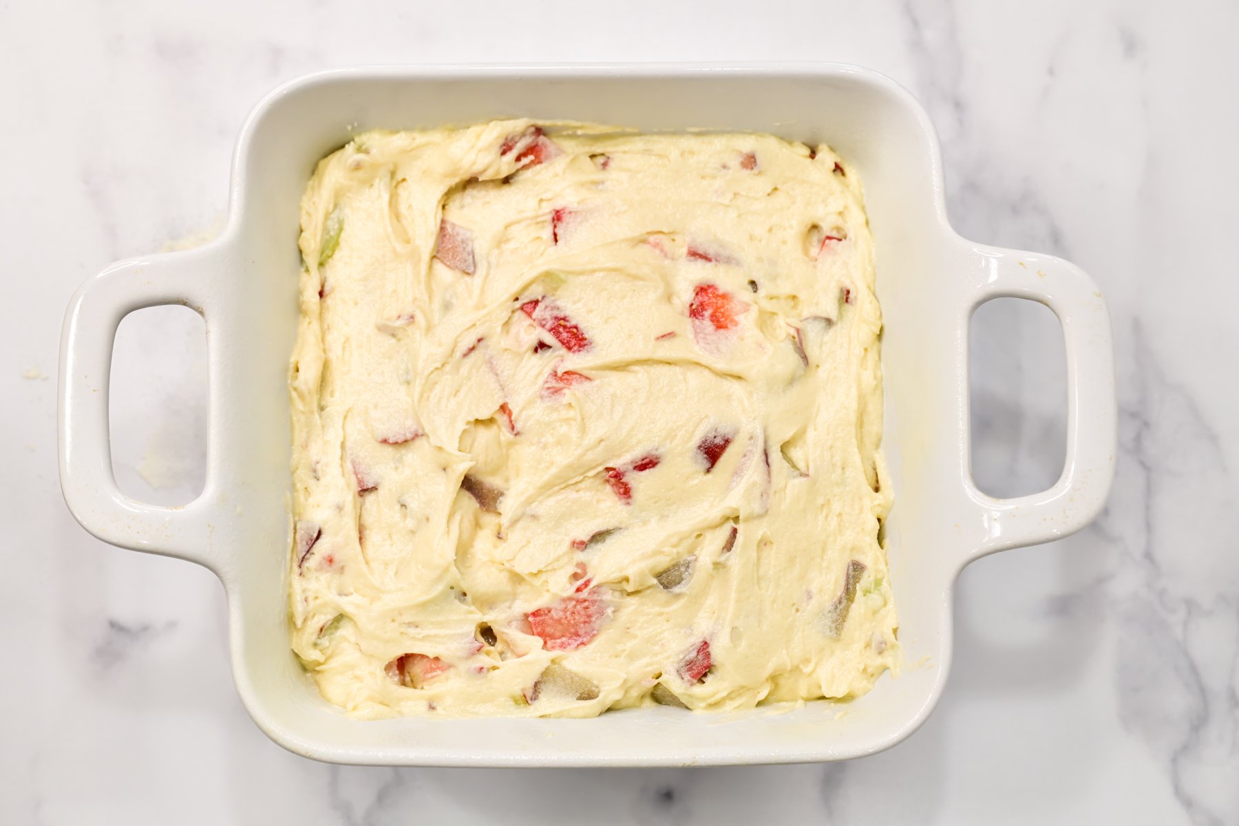 Looking down at a square white baking pan filled with rhubarb coffee cake batter
