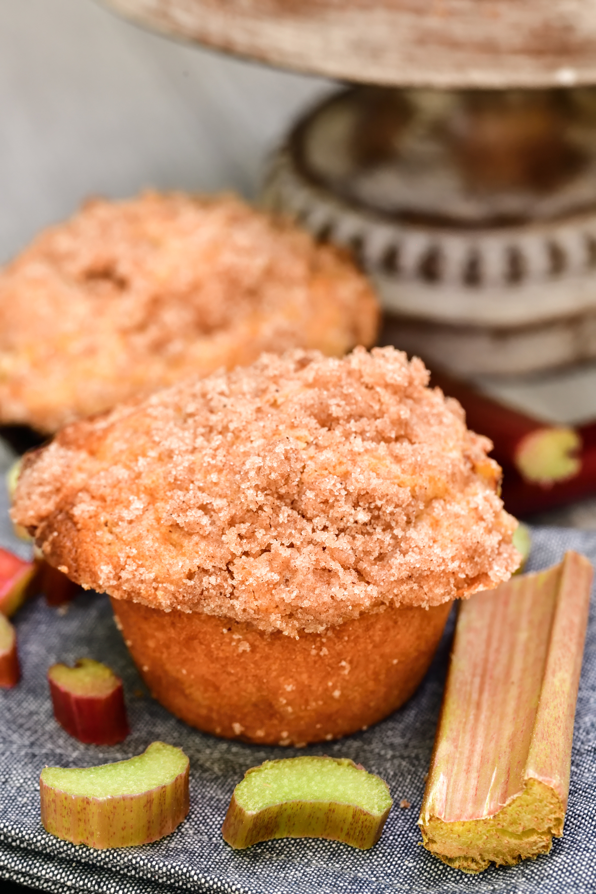 two rhubarb muffins on a grey table cloth surrounded by fresh rhubarb
