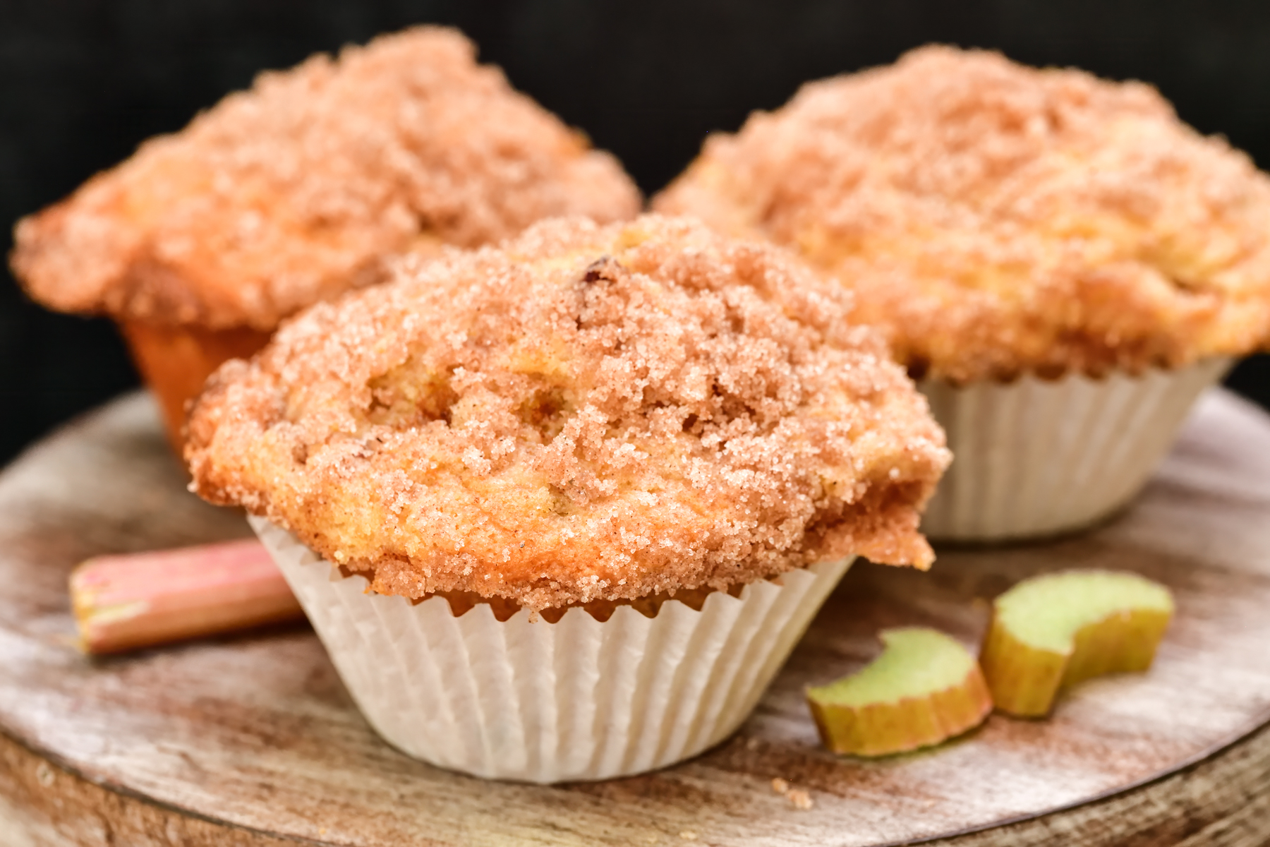 3 Rhubarb muffins with a cinnamon streusel topping on a wood cake stand with sliced rhubarb