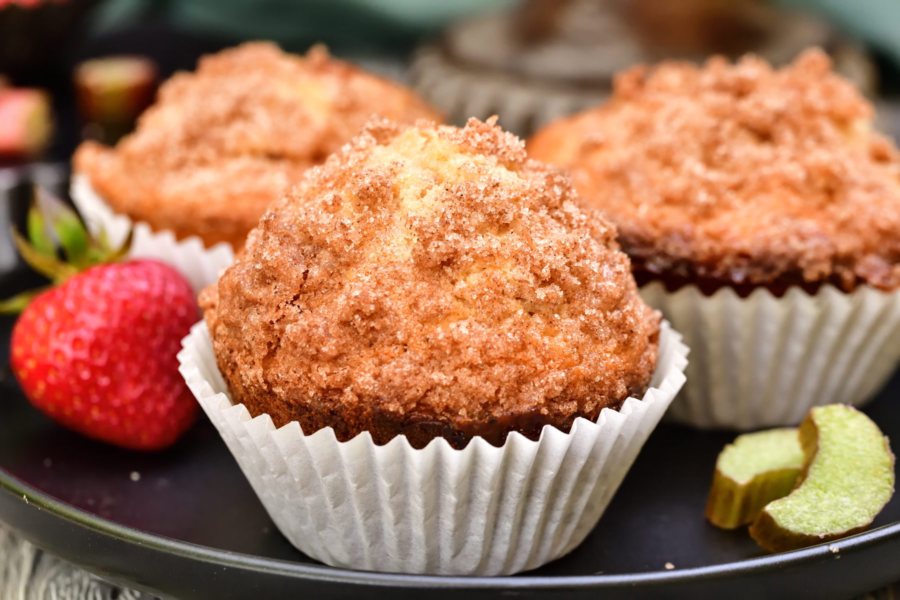 Three Strawberry rhubarb muffins in white paper liners on a black plate with fresh strawberries and rhubarb