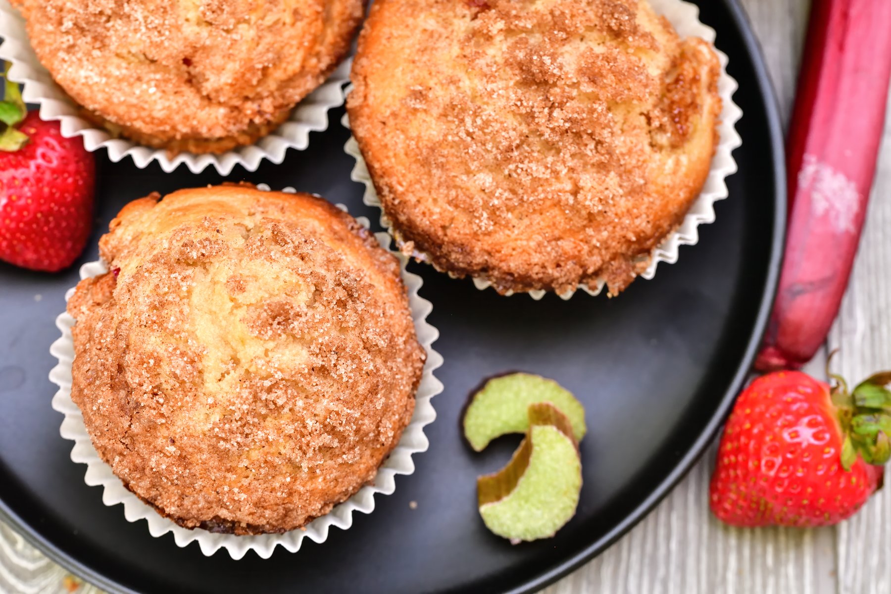 overhead view of 3 muffins on a black plate surrounded by fresh strawberries and rhubarb