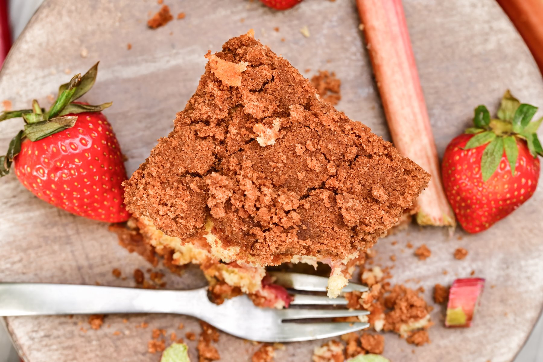 Looking down at a wood plate with a square of cinnamon coffee cake next to strawberries and a stalk of rhubarb and a silver fork